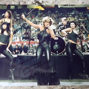 Può includere: Una foto in bianco e nero di una donna che canta sul palco con un microfono. Indossa un abito di pelle nera con una gonna a balze. Altre tre donne sono in piedi sul palco dietro di lei. La foto è firmata "Tina Turner".