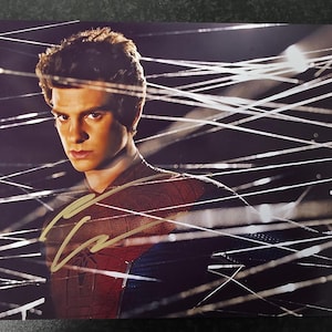 Peut inclure: Portrait d'un homme en costume de Spider-Man, avec une signature à l'encre dorée. L'image a un fond sombre avec des lignes blanches. Le costume est rouge, bleu et noir.