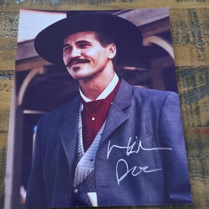 May include: A printed photograph of a man in a dark hat and suit, with a signature in white ink. The man has a mustache and is wearing a dark suit jacket over a vest and a red shirt.