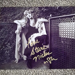 May include: Monochrome photograph of a woman with long, wavy hair, wearing a dark dress and a fringed shawl. The image is signed in gold ink with the name "Stevie Nicks". The woman is leaning against a metal fence.