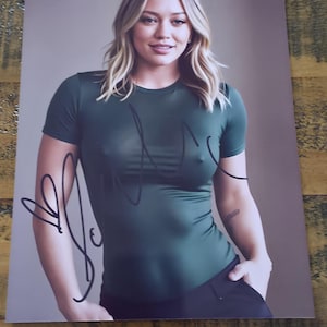 Op de afbeelding: Een foto van een vrouw in een donkergroen shirt met korte mouwen en een zwarte broek. Het shirt heeft een handtekening met zwarte inkt. De vrouw heeft blond haar en glimlacht. De achtergrond is effen en neutraal.