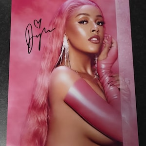 May include: A pink-toned photograph of a woman with pink hair and makeup. She is wearing long, dangling earrings and pink gloves. The image includes an autograph with a heart symbol. The background is a soft pink.