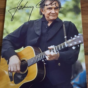Puede incluir: Fotografía firmada de Johnny Cash tocando la guitarra. Viste camisa y chaqueta negras, con una correa para la guitarra. La guitarra es de color marrón claro. La firma es con tinta negra.