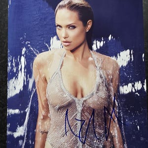 Puede incluir: Una fotografía en blanco y negro de una mujer con el pelo mojado que lleva un top plateado transparente. La mujer mira directamente a la cámara. El fondo es un diseño abstracto azul y blanco. La foto está firmada "Angelina" con tinta azul.