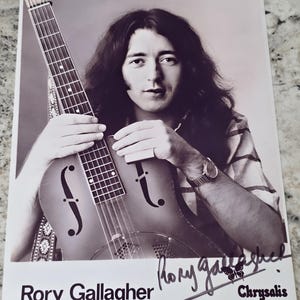 Puede incluir: Fotografía en blanco y negro de Rory Gallagher, un músico irlandés, sosteniendo una guitarra. Lleva una camisa a rayas y tiene el pelo largo. La foto está firmada "Rory Gallagher" y "Chrysalis".
