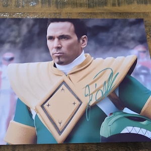 May include: A signed photograph featuring a man in a green and gold costume, likely from a superhero franchise. The costume includes a distinctive helmet and shoulder pads. The image is signed in green ink.