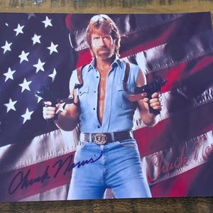 May include: A print featuring a man holding two guns, set against a backdrop of the American flag. The man is wearing a denim shirt and jeans, with a belt buckle. The print is signed "Chuck Norris".