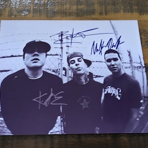 Puede incluir: Fotografía en blanco y negro de tres hombres, probablemente una banda, posando frente a una valla. Cada hombre lleva una camisa oscura y un sombrero. La foto incluye firmas y el logotipo de la banda.