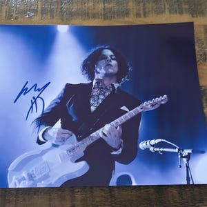 May include: A signed photograph of a musician playing a white electric guitar on stage. The musician is wearing a black suit and a patterned shirt. The image has a blue tint and a signature in the upper left corner.