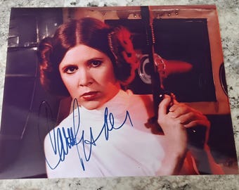 May include: A black and white photo of Carrie Fisher as Princess Leia from Star Wars, signed in blue ink. The photo shows her holding a blaster and looking directly at the camera.