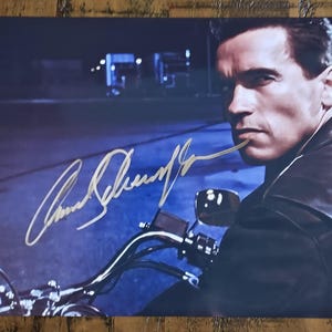 Peut inclure: Photographie d'un homme sur une moto, avec une signature dorée. L'homme porte une veste en cuir noire et regarde sur le côté. L'arrière-plan est bleu foncé avec des lampadaires.