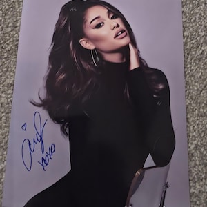Puede incluir: Una fotografía brillante que muestra un retrato de Ariana Grande con cabello oscuro largo, vistiendo una blusa negra de cuello alto de manga larga y aretes de aro. La imagen incluye una firma en tinta azul con un pequeño corazón y el texto xoxo.