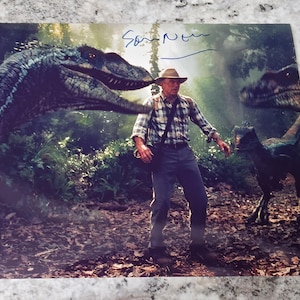 Könnte beinhalten: Ein Farbfoto eines Mannes in einer Dschungelkulisse mit zwei Dinosauriern. Der Mann trägt ein kariertes Hemd und einen Hut. Die Dinosaurier sind ein grün-blauer Velociraptor und ein dunkelgrauer Velociraptor. Der Mann hält eine Waffe. Das Bild ist signiert mit "Sam Neill".