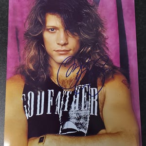 Op de afbeelding: Een foto van een man met lang, golvend haar, gekleed in een zwarte tanktop met het woord "GODFATHER" erop gedrukt. De afbeelding is gesigneerd met blauwe inkt. De achtergrond is een roze en paarse gradiënt.