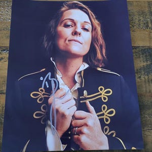 May include: A signed photograph featuring a person in a dark blue military-style jacket with gold embellishments. The jacket has decorative gold trim and buttons. The person is wearing a white collared shirt. The background is a dark, solid color.