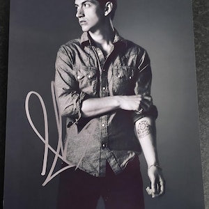 Puede incluir: Fotografía en blanco y negro de una persona con una camisa vaquera y pantalones oscuros, con una firma en la esquina inferior izquierda. La persona tiene un tatuaje en el brazo y mira hacia un lado.