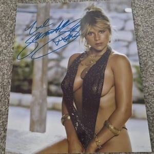 May include: A signed photograph of a woman with blonde hair, wearing a black halter top and gold jewellery. The photo has a blue signature in the upper left corner. The background is a blurred outdoor scene.