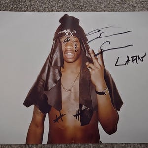 Puede incluir: Fotografía de una persona con un gorro negro con el texto "HOMME de XX", una cofia marrón y un collar dorado. La persona está haciendo el signo de la paz con los dedos. La fotografía está firmada "S. LAPIN".