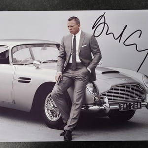Puede incluir: Fotografía de un hombre con traje gris posando junto a un coche plateado. El coche tiene un diseño clásico con una prominente parrilla delantera. La imagen incluye una firma con tinta negra.