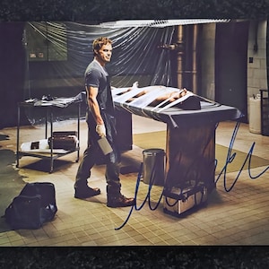 May include: A printed photograph featuring a man in a dark t-shirt and jeans, holding a cleaver, standing near a table with a person strapped to it. The scene is set in a dimly lit room with medical equipment and a signature.