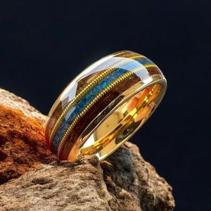 May include: A gold ring with a brown wood inlay and blue accents. The ring is sitting on a piece of wood.