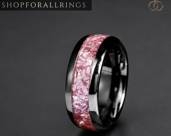 May include: A black ring featuring a central inlay of crushed pink stone fragments. The band has a polished, reflective finish. The text SHOPFORALLRINGS is visible in the upper left corner, and a small crown and ring logo appears in the upper right.