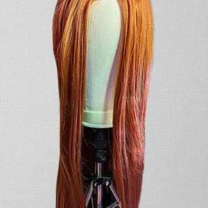 May include: A long, straight wig with a blend of orange and reddish-pink tones. The wig is styled on a mannequin head, showcasing its length and color variations. The hair is parted down the middle, and the wig is secured on a black tripod stand.