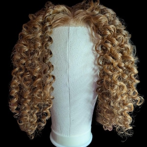 May include: A shoulder-length wig featuring light brown, golden-toned hair with tight, defined ringlet curls. The hair is parted in the center and styled on a beige canvas mannequin head, showcasing a lace-like front hairline construction.