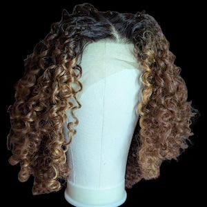May include: A shoulder-length wig featuring tight, defined curls with a dark brown base color transitioning into lighter golden-brown highlights. The wig is displayed on a white mannequin head and includes a sheer lace front hairline.