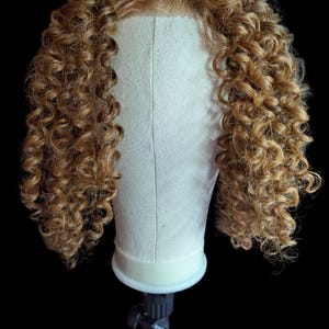 May include: A light brown curly wig on a canvas head form. The wig features tight, voluminous curls and a natural-looking hairline. The head form is mounted on a black stand against a black background, highlighting the wig's texture and color.
