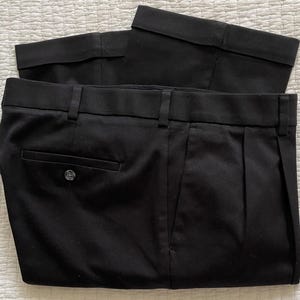 May include: Black dress pants with a buttoned back pocket and belt loops. The pants are folded, showing the waistband and pleats. The fabric appears to be a solid, dark color.