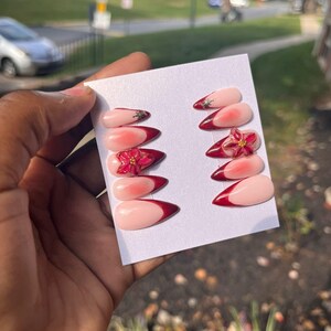 May include: A set of press-on nails with a light pink base and a deep red French tip. Some nails feature a floral design with gold accents. The nails are displayed on a white card.