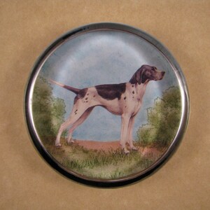 May include: A round glass paperweight with a silver rim features a painted image of a white and brown dog with black spots standing in a grassy field.