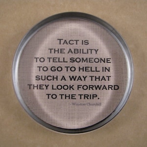 May include: A round, silver-colored coaster with a black quote that reads "Tact is the ability to tell someone to go to hell in such a way that they look forward to the trip." The quote is attributed to Winston Churchill.