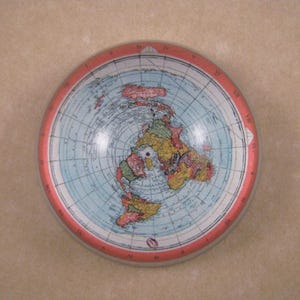 May include: A round, vintage-style map of the world, featuring a red border and a light blue background. The map shows the continents in shades of yellow, green, and red. The design is a flat-earth projection.
