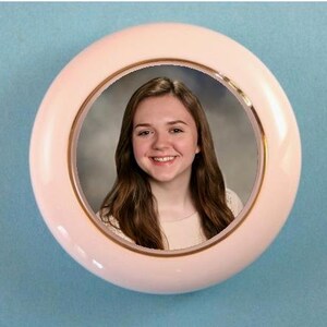 May include: A round, light pink picture frame with a gold-colored inner rim. The frame holds a portrait of a smiling person with long brown hair. The background is a light blue.
