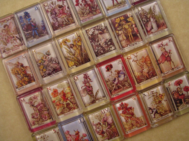 May include: A set of alphabet tiles with colorful illustrations of fairies and flowers. Each tile features a letter of the alphabet and a corresponding image. The tiles are made of glass and have a glossy finish.