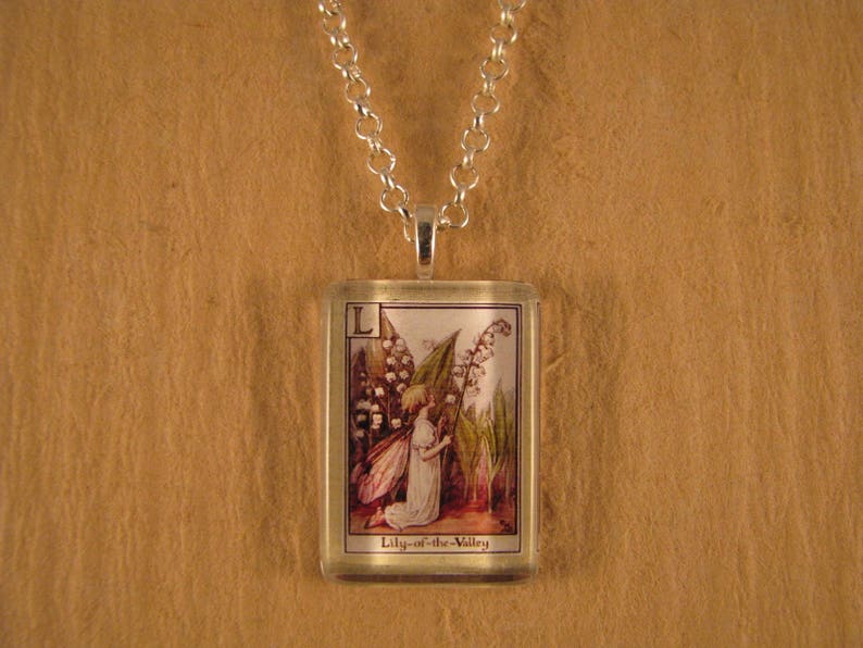 May include: A silver necklace with a rectangular glass pendant featuring a vintage illustration of a fairy with wings and a bouquet of lily-of-the-valley flowers. The pendant has the letter "L" and the text "Lily-of-the-Valley" printed on it.