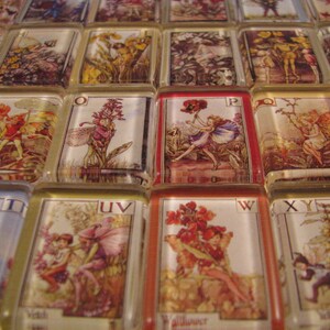 May include: A set of alphabet tiles featuring illustrations of fairies and flowers. Each tile has a letter of the alphabet and a corresponding illustration. The tiles are made of glass and have a gold trim.