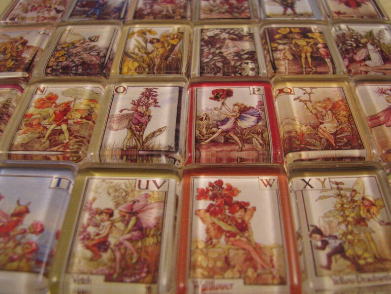 May include: A set of alphabet tiles featuring illustrations of fairies and flowers. Each tile has a letter of the alphabet and a corresponding illustration. The tiles are made of glass and have a gold trim.