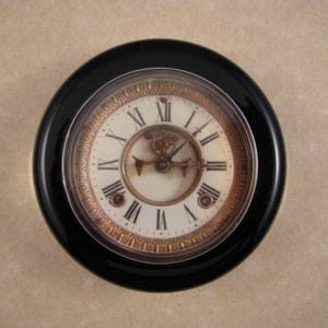 May include: A black and gold clock with Roman numeral markings on a white face. The clock has a visible gear mechanism and is set against a beige background.