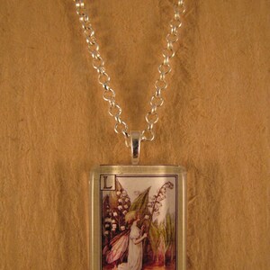 May include: A silver chain necklace with a glass pendant featuring a vintage illustration of a fairy kneeling next to a lily of the valley flower. The pendant has the letter "L" and the text "Lily-of-the-Valley" printed on it.