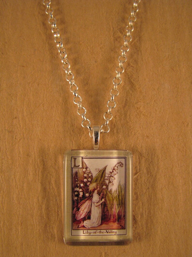 May include: A silver chain necklace with a glass pendant featuring a vintage illustration of a fairy kneeling next to a lily of the valley flower. The pendant has the letter "L" and the text "Lily-of-the-Valley" printed on it.
