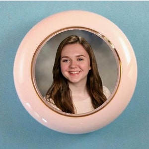 May include: A white ceramic round picture frame with a gold trim. The frame has a photo of a young woman with long brown hair. She is smiling and wearing a white shirt.