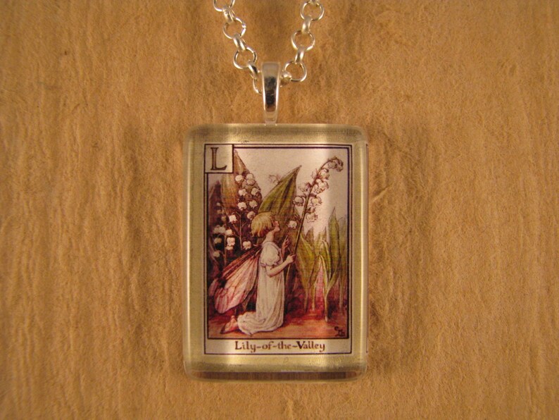 May include: A glass pendant necklace with a silver chain. The pendant features a vintage illustration of a fairy holding a lily-of-the-valley flower. The text "Lily-of-the-Valley" is printed below the illustration.