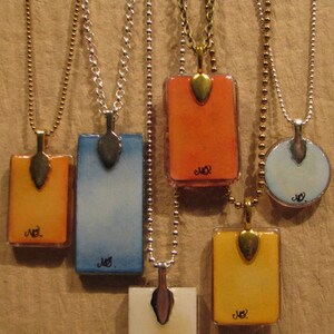 May include: Five necklaces with rectangular glass pendants on silver chains. The pendants are painted with watercolor washes in shades of orange, blue, yellow, and white. Each pendant has a gold leaf accent and the word "Joy" written in black ink.