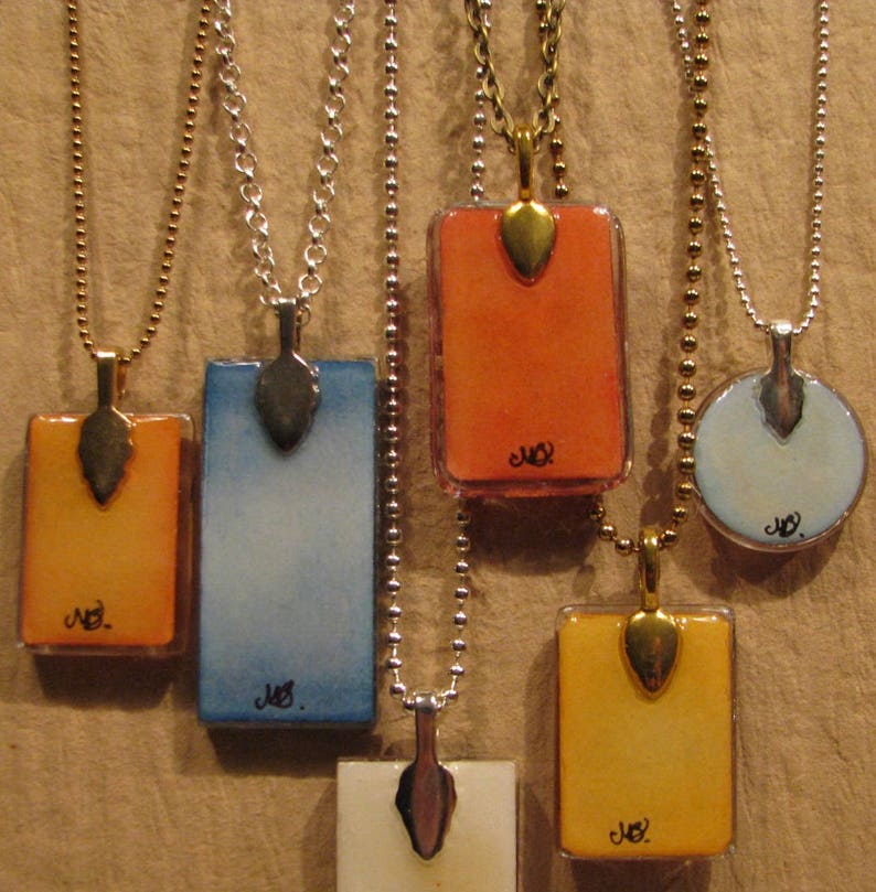 May include: Five necklaces with rectangular glass pendants on silver chains. The pendants are painted with watercolor washes in shades of orange, blue, yellow, and white. Each pendant has a gold leaf accent and the word "Joy" written in black ink.