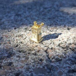 Op de afbeelding: Een gouden metalen bedel in de vorm van een klein gebouw met een puntdak en een decoratief ontwerp. De bedel ligt op een ruw, grijs oppervlak.