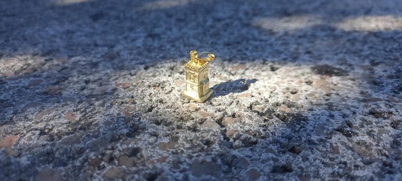 Op de afbeelding: Een gouden metalen bedel in de vorm van een klein gebouw met een puntdak en een decoratief ontwerp. De bedel ligt op een ruw, grijs oppervlak.