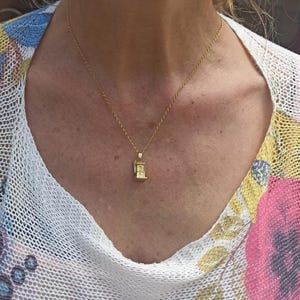 Op de afbeelding: Een gouden ketting met een klein gouden hanger in de vorm van een gebouw. De hanger heeft een gedetailleerd ontwerp en hangt aan de borst van een vrouw.